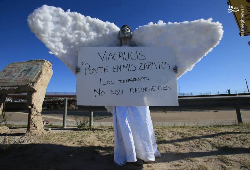 مردی با لباس فرشته تابلویی در دست دارد که روی آن نوشته شده است: "در کفش من باش. مهاجران جنایتکار نیستند" در چند متری حصار مرزی بین مکزیک و ایالات متحده